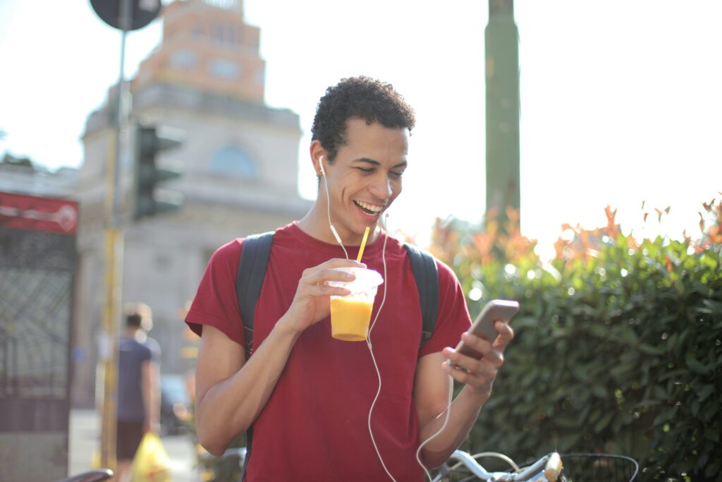 A man using mobile while walking in public and having a juice glass in his hands.