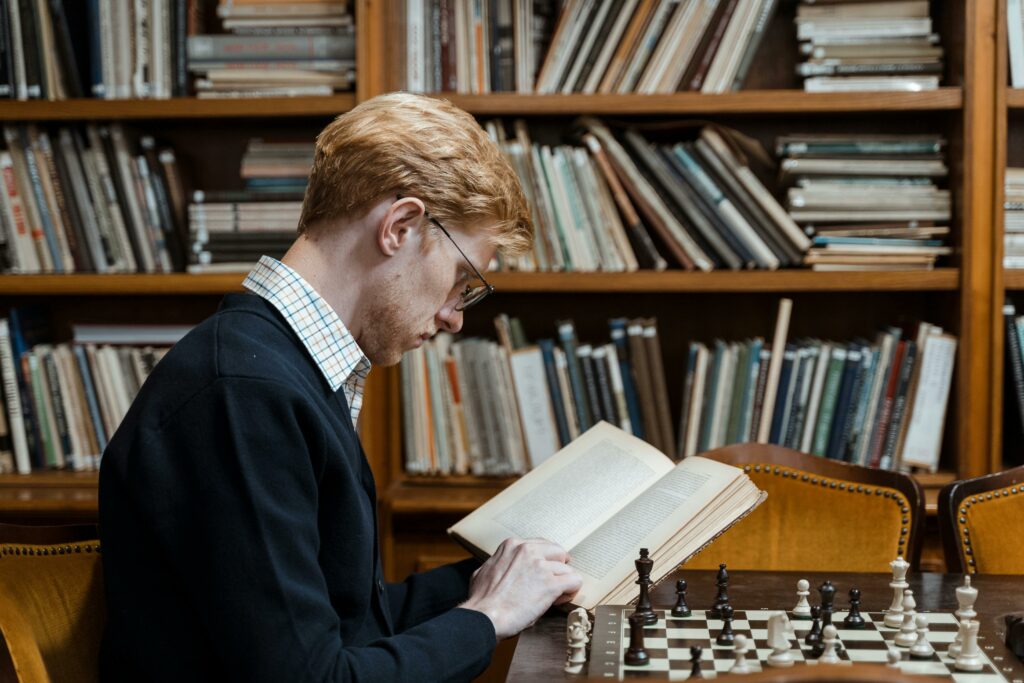 A man is reading book While having a chess in front of him.